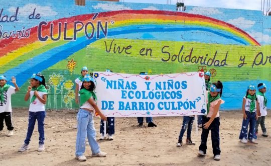 Children in green kerchiefs hold a banner that reads, "Environmentment Boys and Girls of Culpón" in front of a colourful mural.